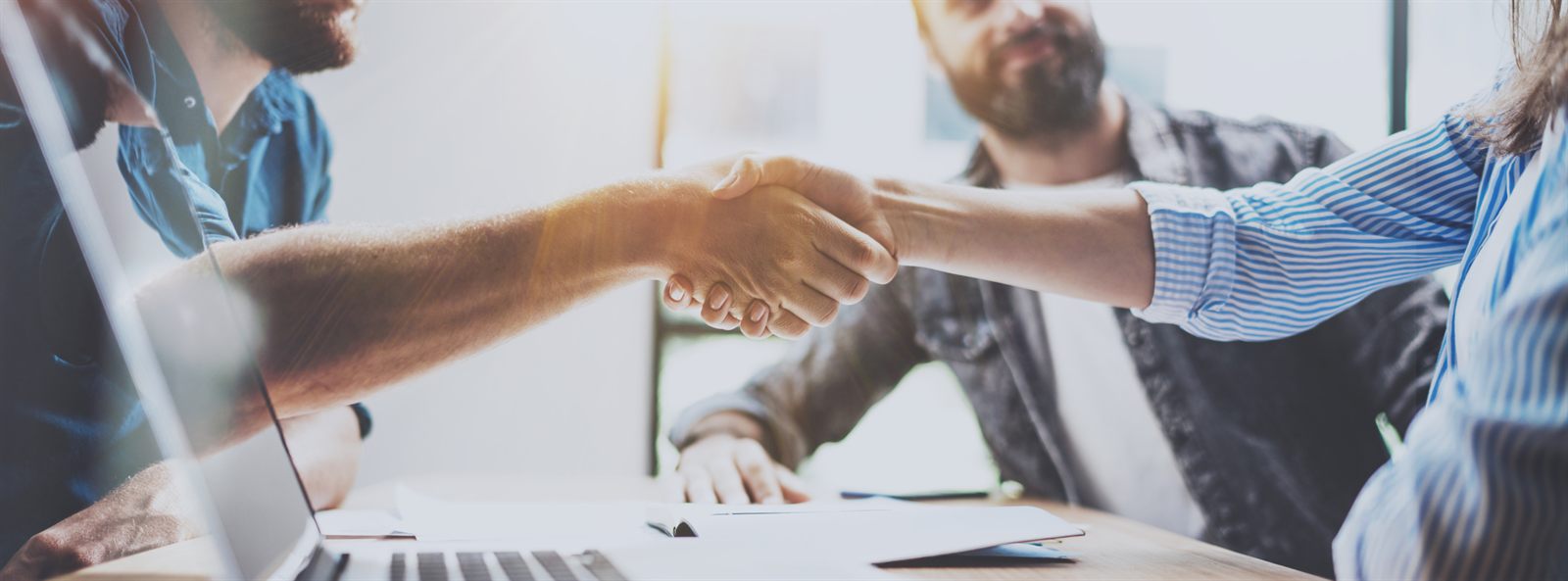Two people shaking hands across a desk with a laptop